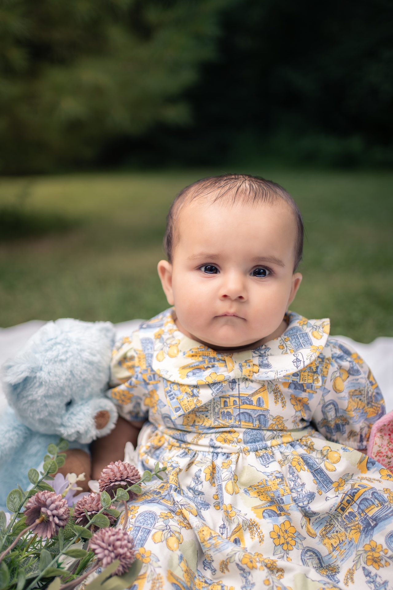 A baby in a lemon-and-blue printed dress sits on a blanket and looks straight at the camera.