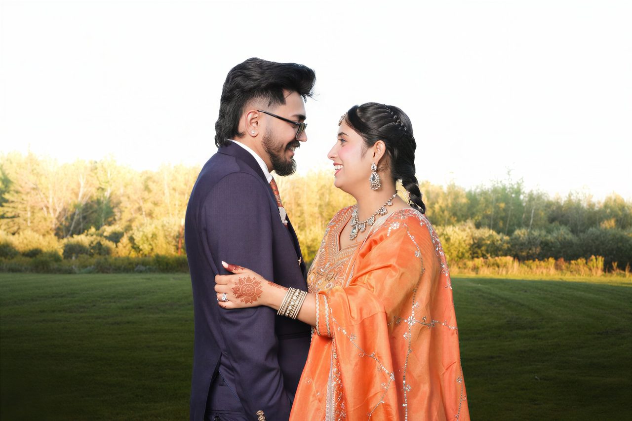 A couple stands close and smiles at each other in a grassy field in warm evening light, she in an embroidered orange outfit and he in a navy suit.