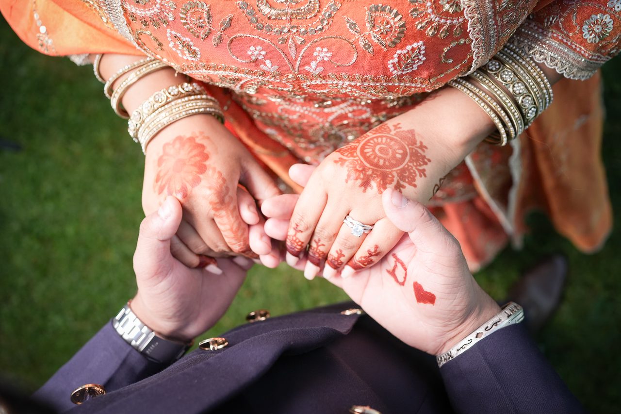 Seen from above, a couple holds hands on the grass; one hand shows a floral henna design and a ring, beside gold bangles and an embroidered orange sleeve.