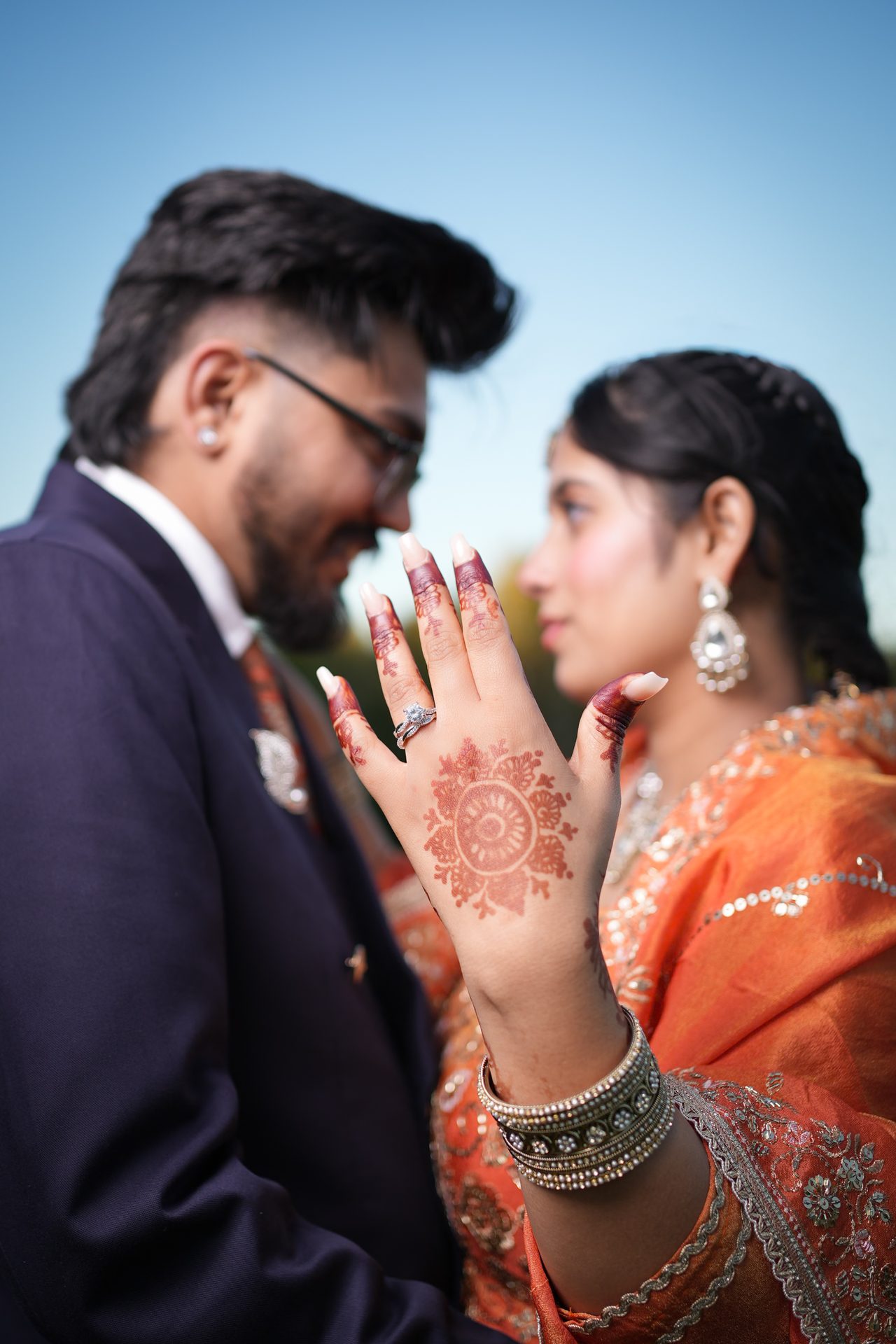 A hand with a henna design and a diamond ring is raised in sharp focus while a couple smiles at each other, softly blurred, behind it.