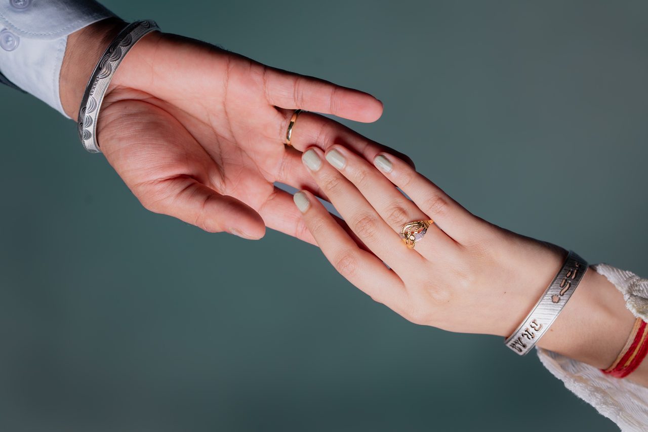 Two hands meet against a teal background: one open palm wearing a silver bangle and a gold band, the other resting on its fingers with a gold ring, pale green nails and a silver bracelet.