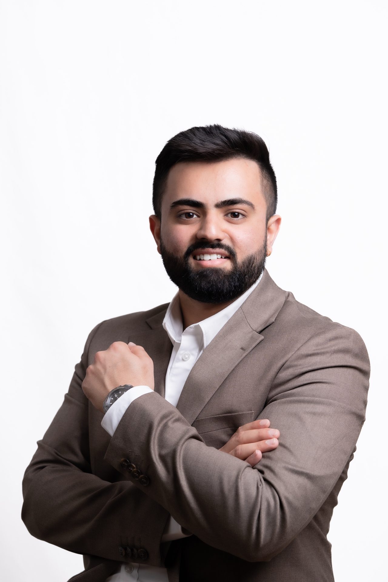 Studio portrait of a smiling bearded man in a brown blazer with his arms crossed, on a white background.