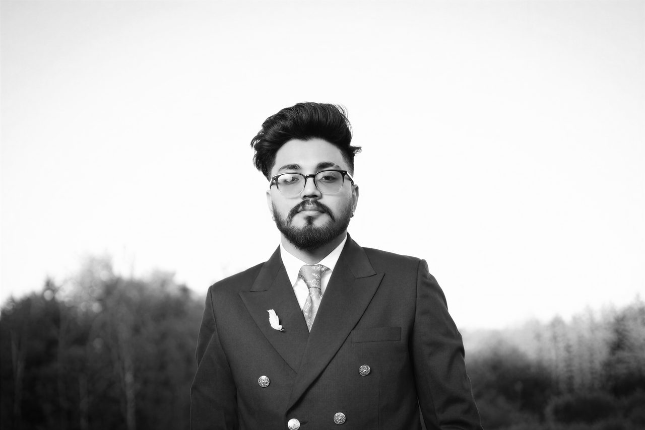 Black-and-white portrait of a bearded man in glasses and a double-breasted suit, looking at the camera against a bright sky.