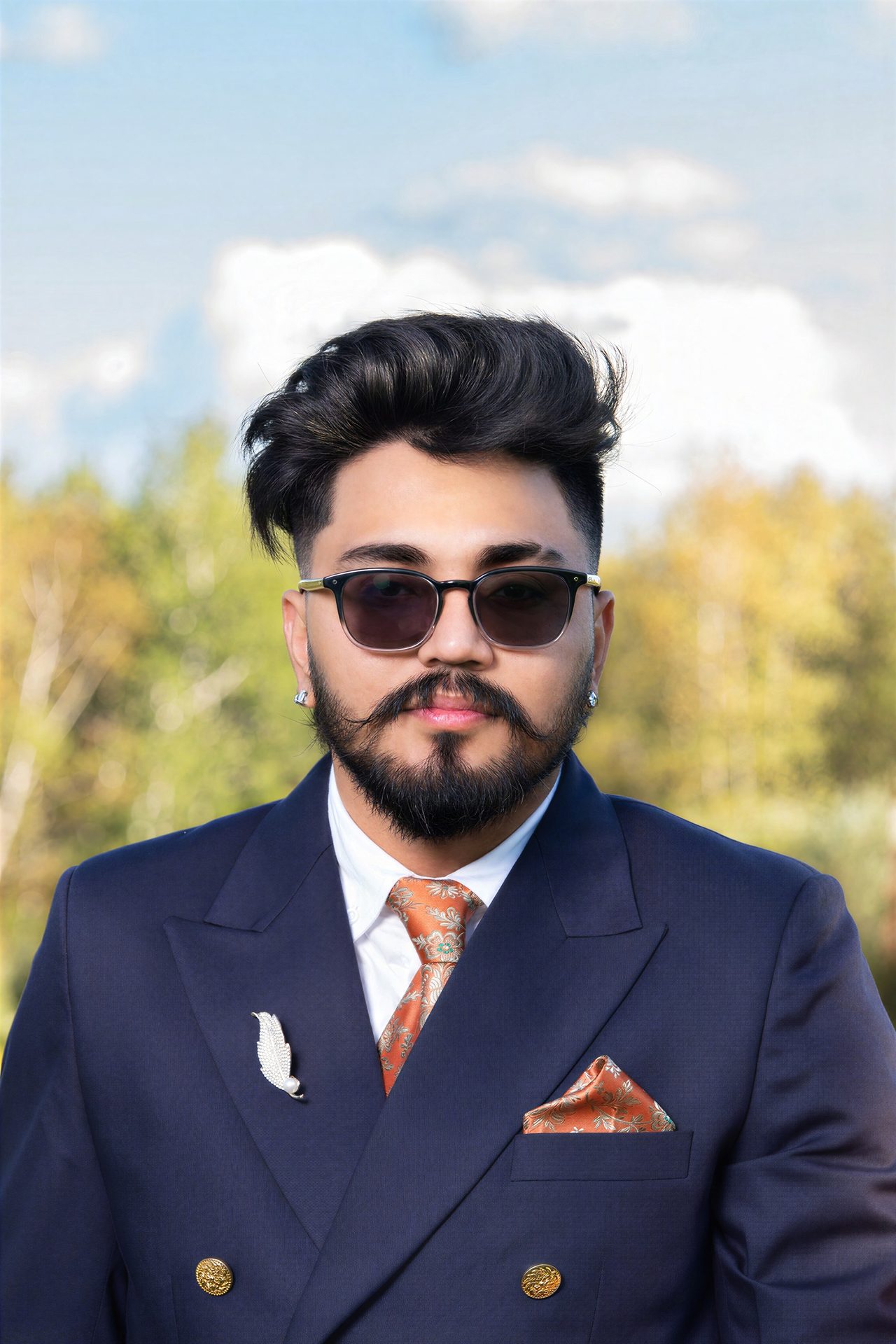 A bearded man in sunglasses and a navy suit with an orange tie, against a blue sky and trees.