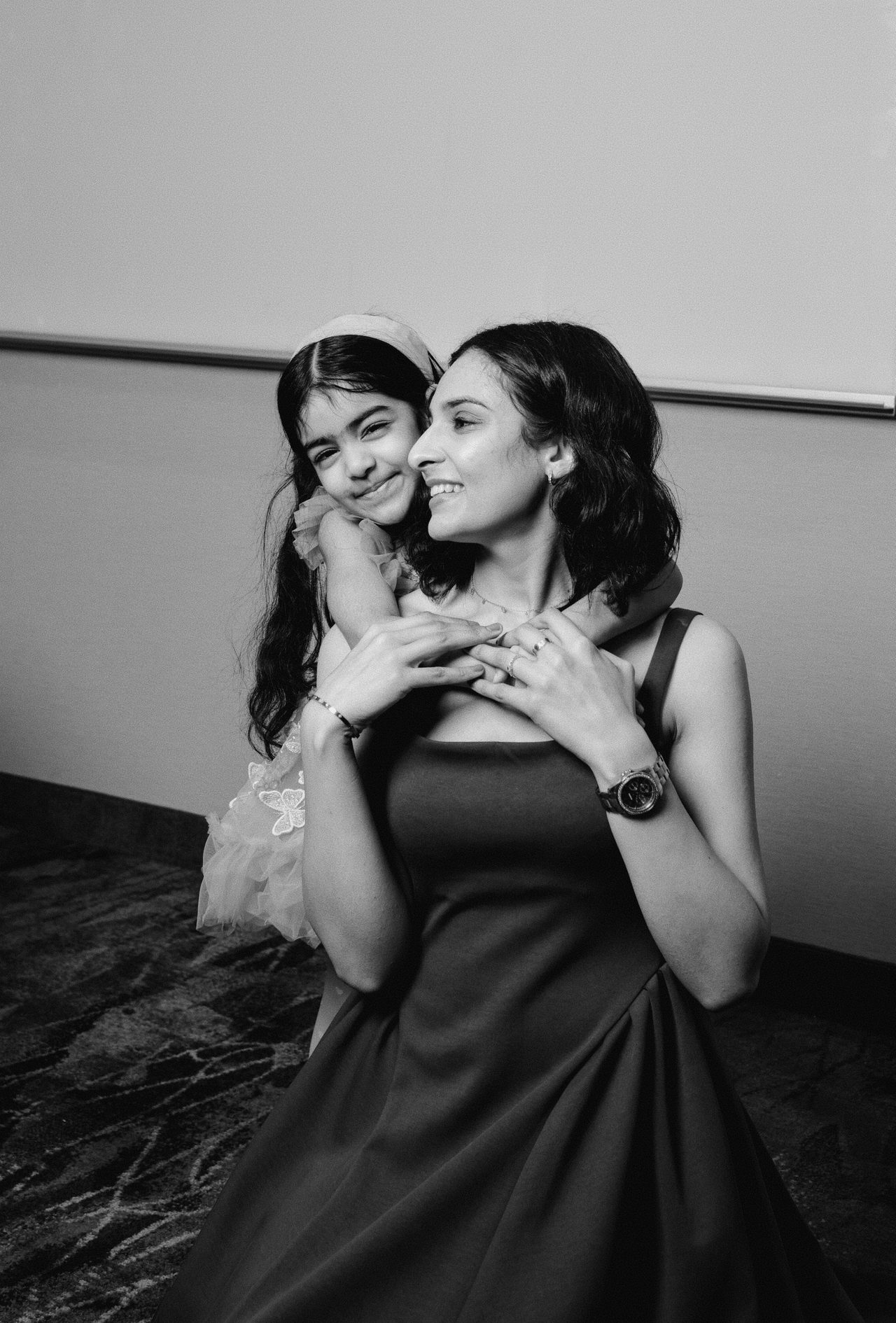 Black-and-white portrait of a woman in a dress smiling off camera as a young girl hugs her from behind.