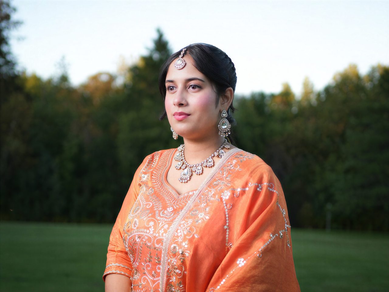A woman in an embroidered orange outfit and traditional jewellery looks into the distance, dark trees behind her at dusk.