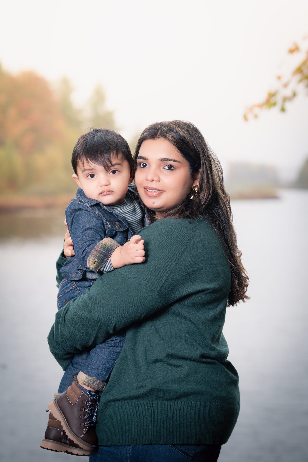 A woman in a green sweater holds a toddler in a denim jacket in front of a misty autumn lake.